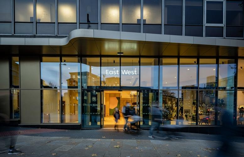 Exterior entrance of the EastWest building on Tollhouse Hill featuring a glass facade and warm lighting.