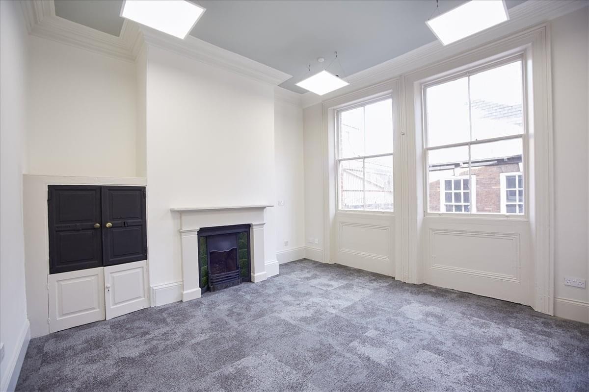 Bright, empty office space with a white fireplace, grey carpet, and large sash windows.