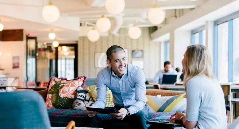 Bright communal lounge with colorful pillows, globe pendant lights, and people chatting.