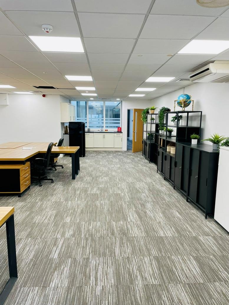 Open-plan office with grey carpet, wooden desks, and black shelving units adorned with green plants.
