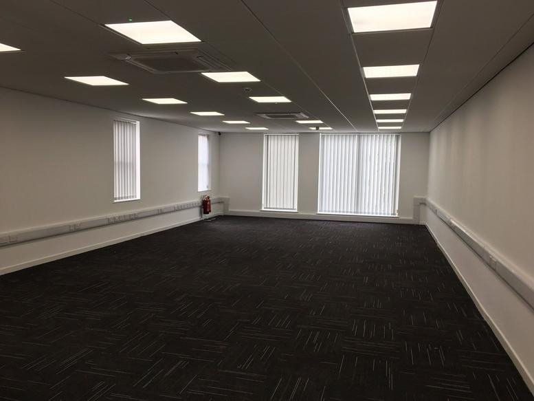 Spacious, empty open-plan office with dark carpeting and bright overhead panel lighting.