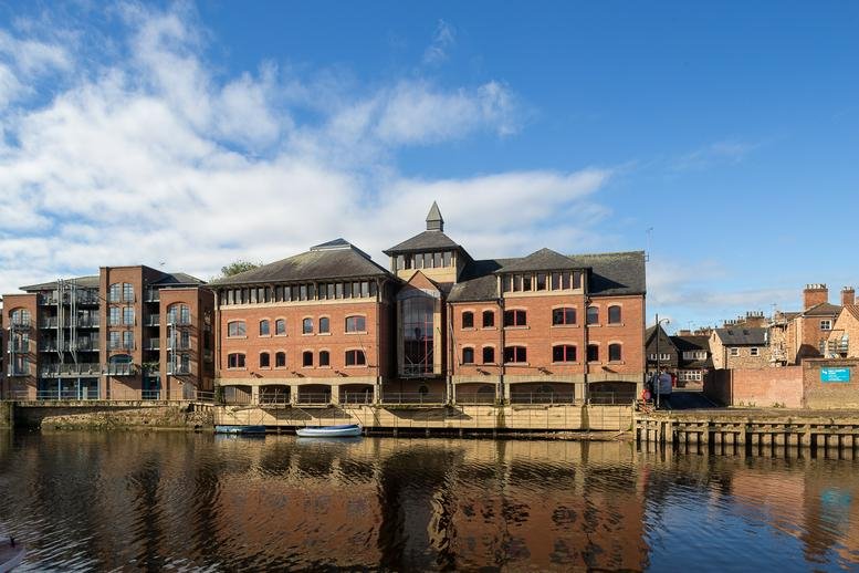 Wide exterior of the brick office building reflected in the river water under a blue sky.