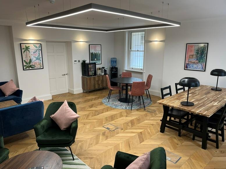 Bright communal lounge featuring velvet armchairs, wooden herringbone floors, and a modern square pendant light.