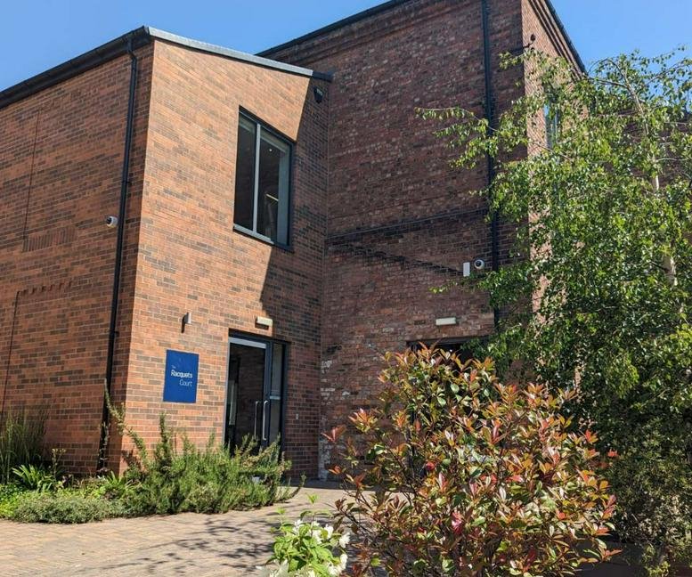 Exterior brick facade and entrance of The Racquets Court with green shrubbery.