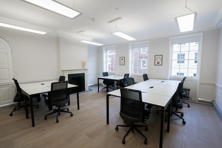 Spacious shared workspace with several white desks and large windows overlooking the street.