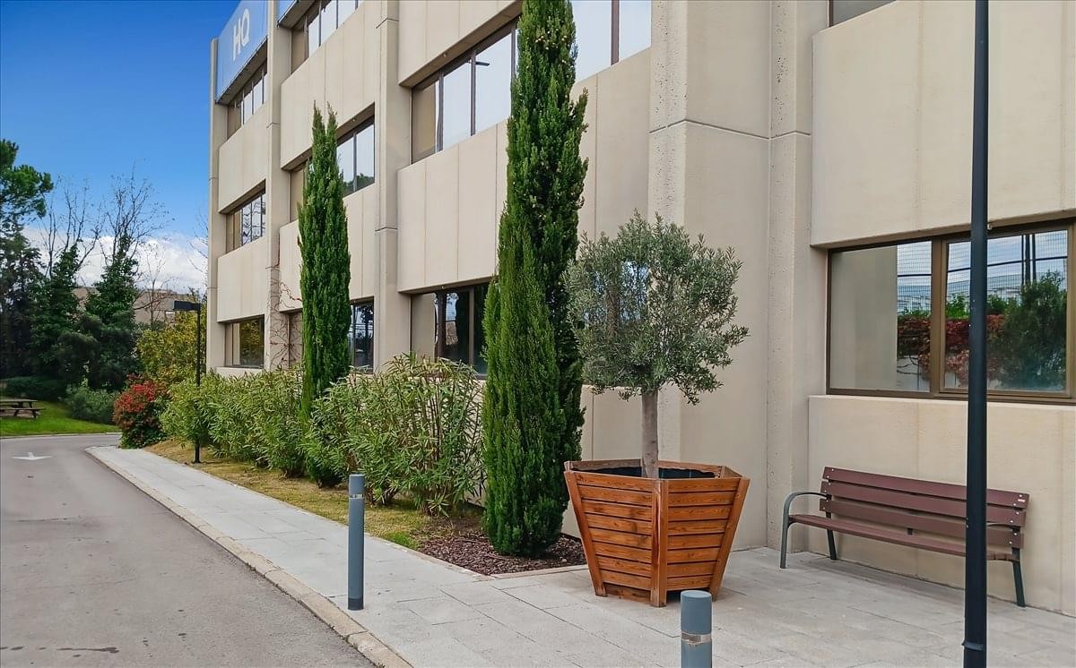 Exterior view of the modern tan building at 35 Calle De Gobelas, Madrid featuring manicured trees and a bench.