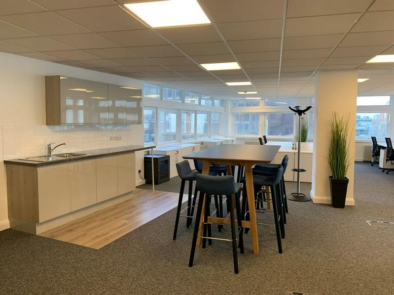Communal breakout space featuring a high wooden table with blue stools and a kitchenette.
