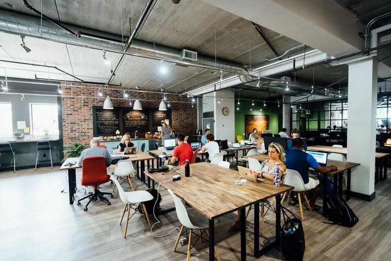 Bustling coworking space with professionals at wooden desks under exposed industrial ceilings.