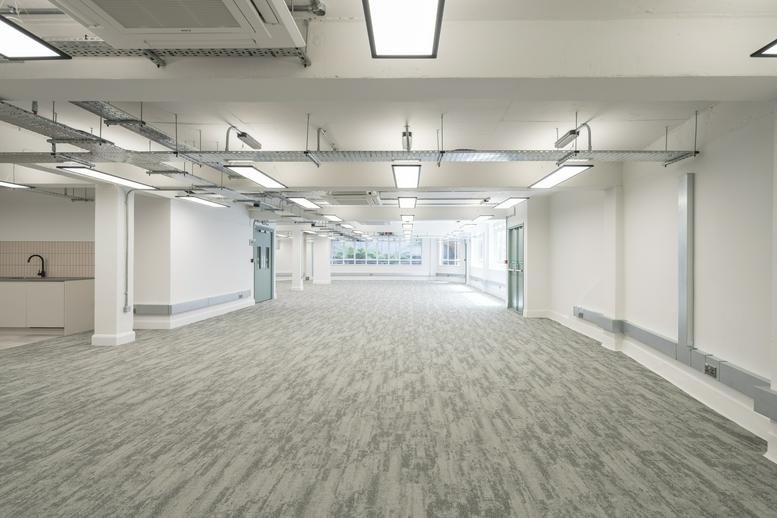Spacious open-plan office area with grey patterned carpeting and a small kitchen unit in the background.