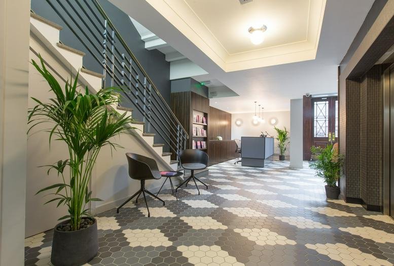 Bright lobby with a geometric patterned floor, a black metal staircase, and lush green potted plants.