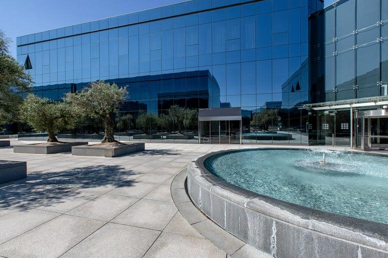 Modern glass exterior of Business Park La Finca with a circular fountain and olive trees in the foreground.