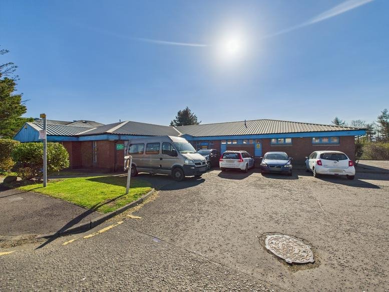 Wide shot of the building entrance featuring a parked silver van and several cars in the lot.