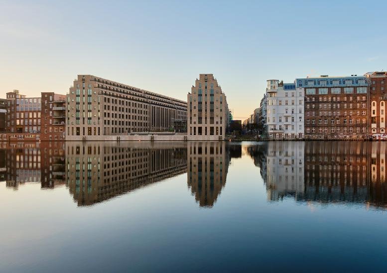 Exterior view of the stepped-facade buildings at Cuvrystraße 53 reflected in the river at dusk.