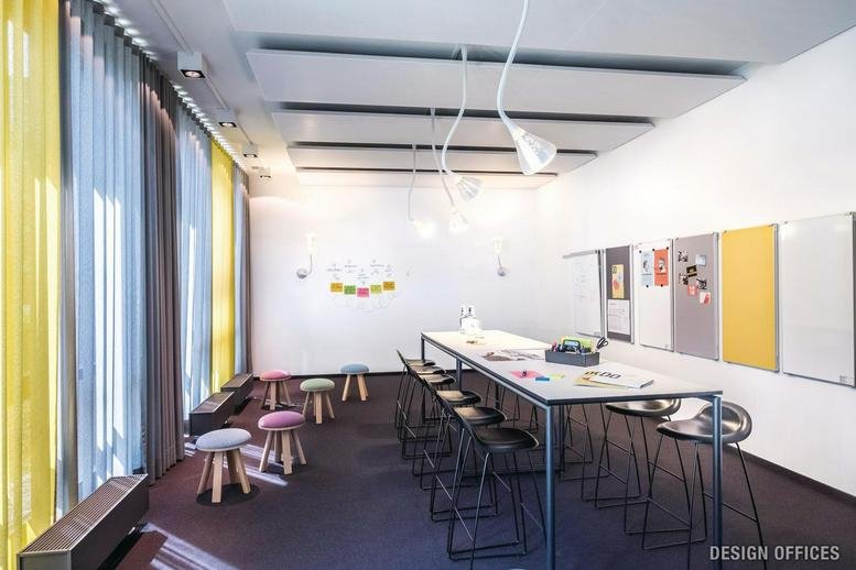 Bright breakout space at Design Offices München Arnulfbogen with colorful stools and long tables.