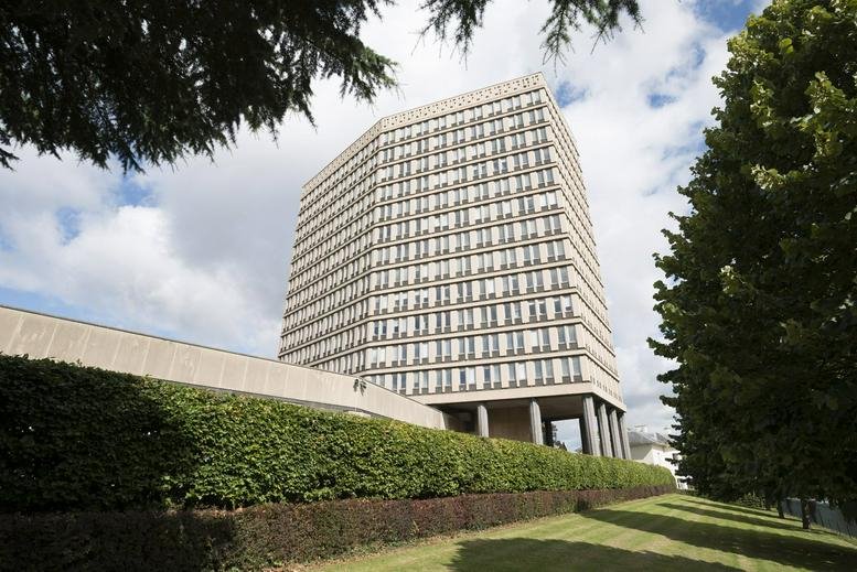 Modern high-rise office building with a light-colored facade and many windows behind a green hedge.