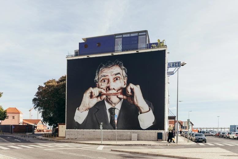 Exterior of the Edifício LACS, Rocha Conde de Óbidos, featuring a large street art portrait on the facade.