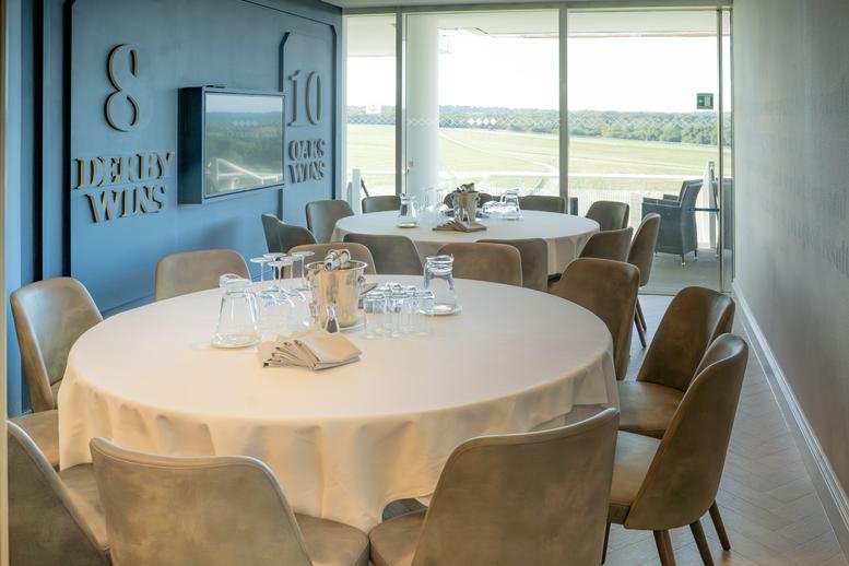 Hospitality suite at Epsom Downs Racecourse featuring round tables, grey chairs, and racetrack views.