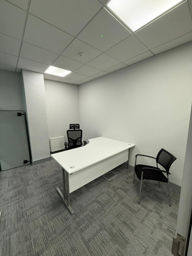 Private single-person office with a large white L-shaped desk and grey textured carpeting.