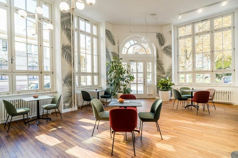 Bright communal lounge with leaf-patterned wallpaper, green and red chairs, and large arched windows.