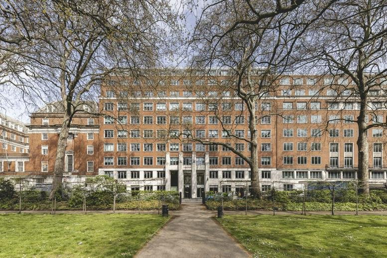 Wide exterior view of the large brick building behind a green park with mature trees.
