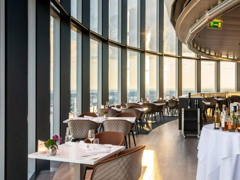 Sunlit dining area with curved windows overlooking the city and set white-clothed tables.