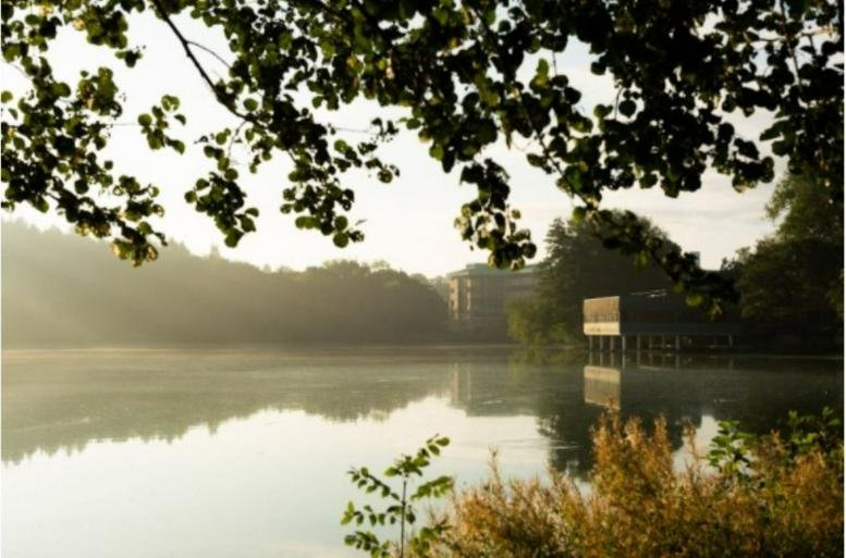 Lakeside view of the No. 1 Mereside, Alderley Park building surrounded by lush greenery at sunrise.