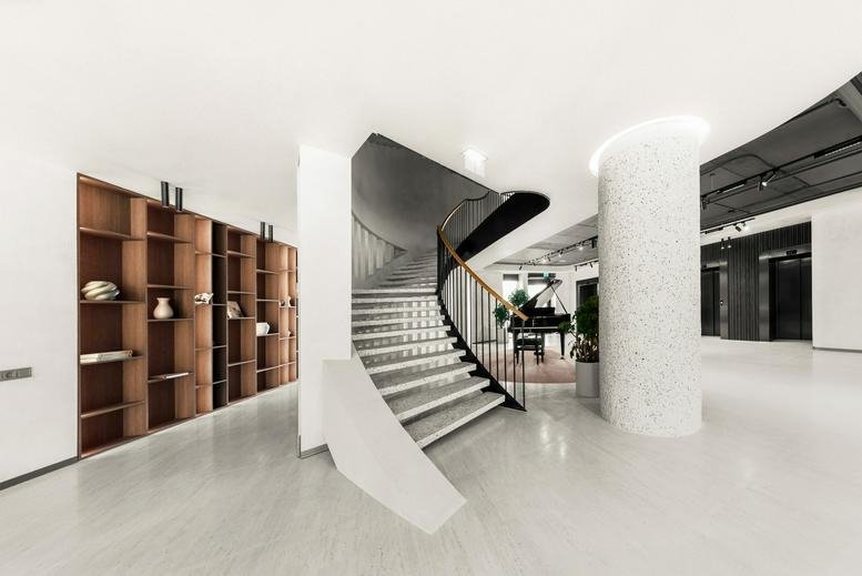 Elegant white spiral staircase next to a minimalist wooden shelving unit in a bright lobby.