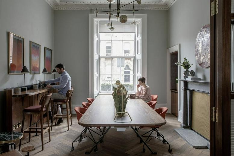 Bright coworking lounge with a long wooden communal table, herringbone floors, and large window.