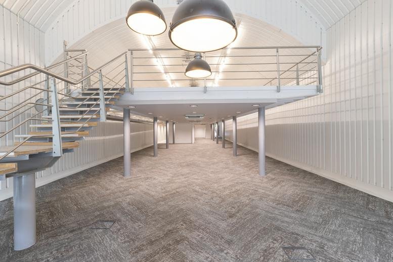 Spacious empty office floor under a mezzanine with industrial lighting and grey carpet.