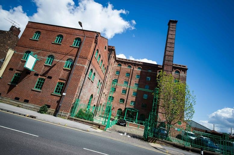 Exterior view of the historic red brick Aizlewood Mill with its tall chimney against a blue sky.