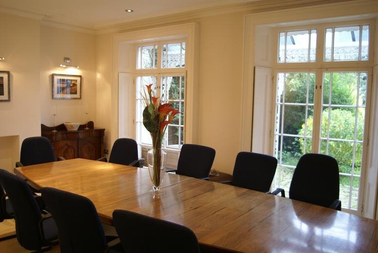 Bright boardroom with a large wooden table, black chairs, and tall windows overlooking greenery.