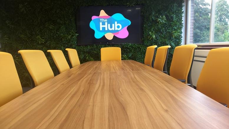 Professional meeting room featuring a long wooden table, yellow chairs, and a green feature wall with a TV screen.