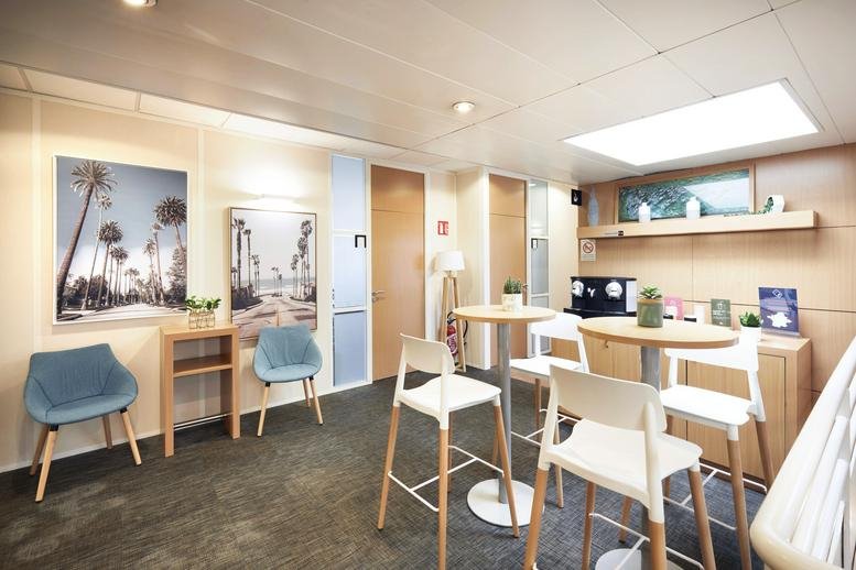 Communal breakout area at Europarc de Pichaury with high-top tables, blue armchairs, and tropical wall art.