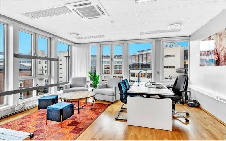 Spacious private office at Hoffsveien 1 featuring large windows, wood floors, and a lounge area with blue ottomans.