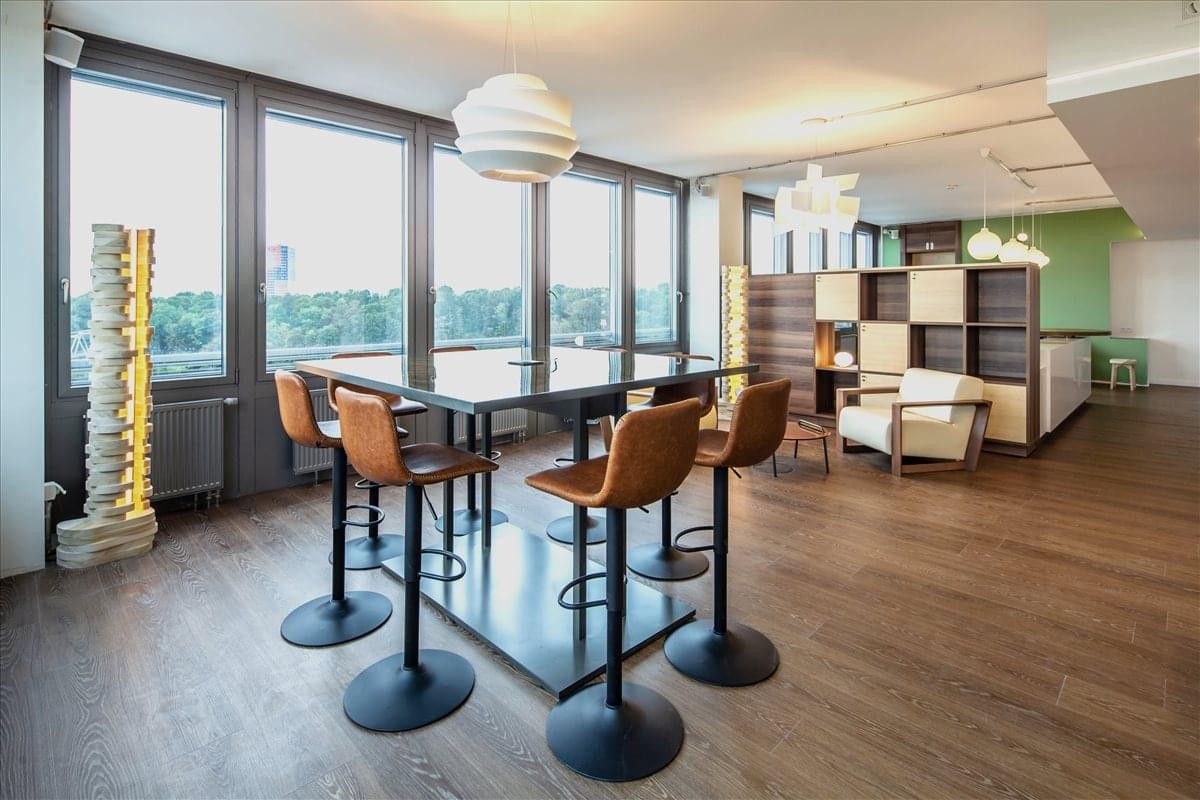Spacious communal breakout area with high tables, armchairs, and floor-to-ceiling windows.