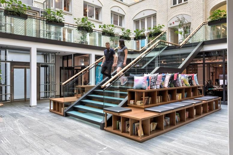 Grand interior courtyard at Krausenstraße 9 with a glass staircase, tiered wooden seating, and hanging plants.
