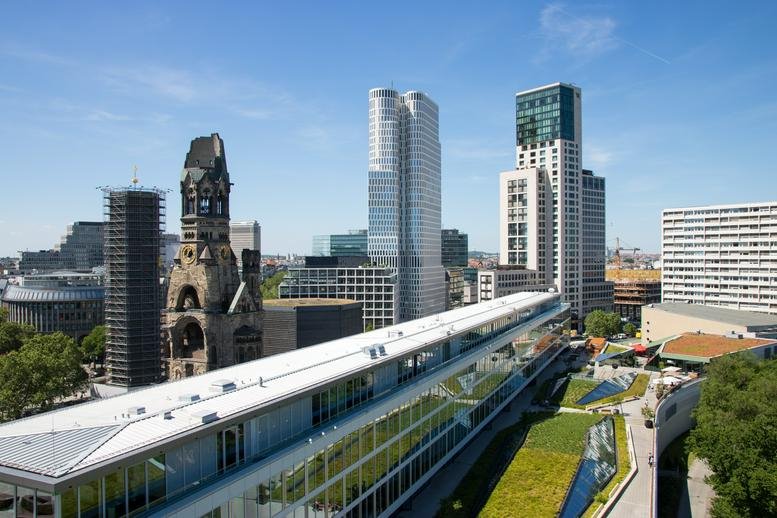 Exterior view of the Kurfürstendamm 11 building featuring the Kaiser Wilhelm Memorial Church and modern skyscrapers.