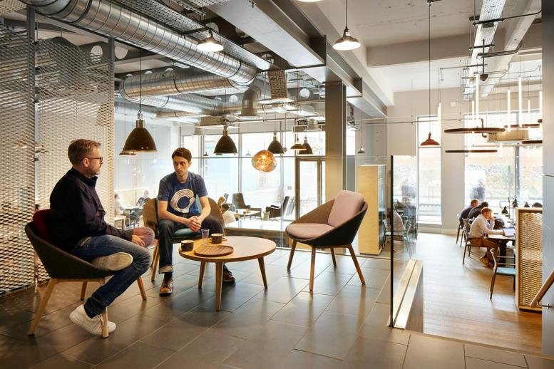 Modern breakout area with industrial ceilings, designer seating, and two people conversing.