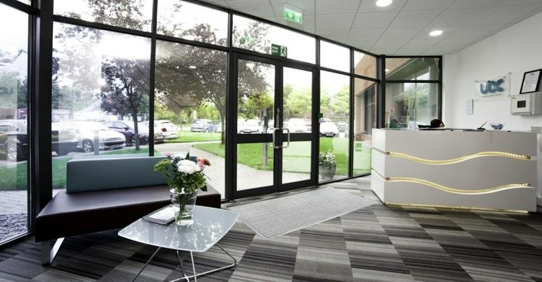 Bright reception lobby featuring a patterned grey carpet, glass entrance doors, and a sleek front desk.