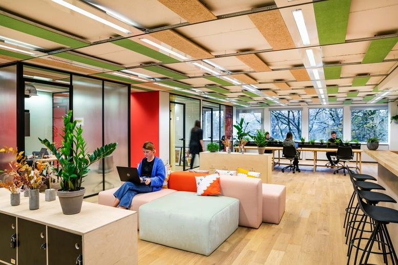 Open-plan coworking lounge with modular pastel seating, plants, and colorful ceiling panels.