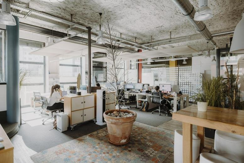Large coworking area with white desks, indoor plants, and exposed concrete ceilings.