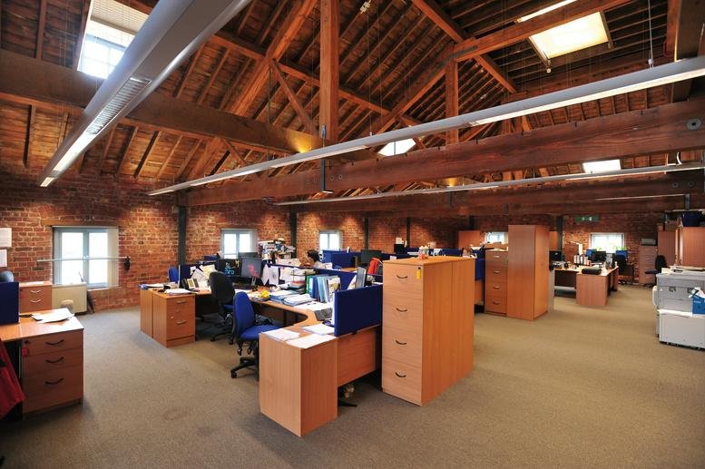 Open-plan office with wooden ceiling beams, brick walls, and multiple workstations with filing cabinets.