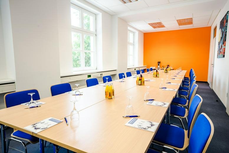 Meeting room at Zur Wetterwarte 50 / Hugo-Junkers-Ring 1-9 with a long table, blue chairs, and an orange feature wall.
