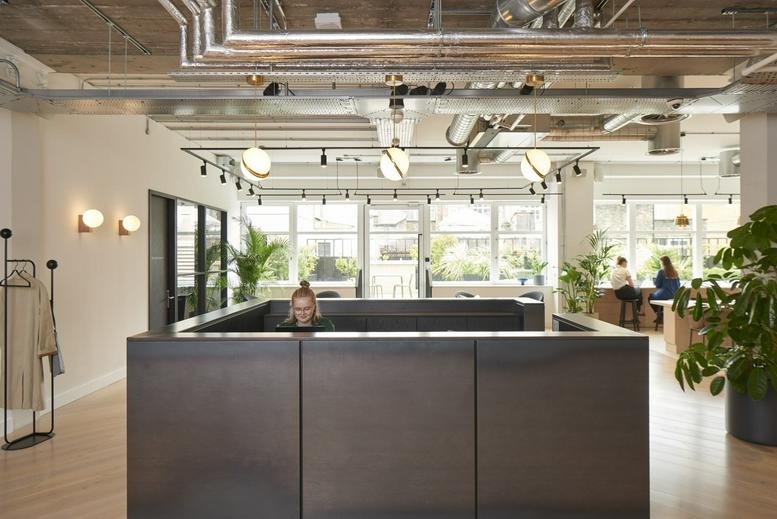 Bright reception area featuring a dark metal desk, indoor plants, and exposed ceiling pipes.