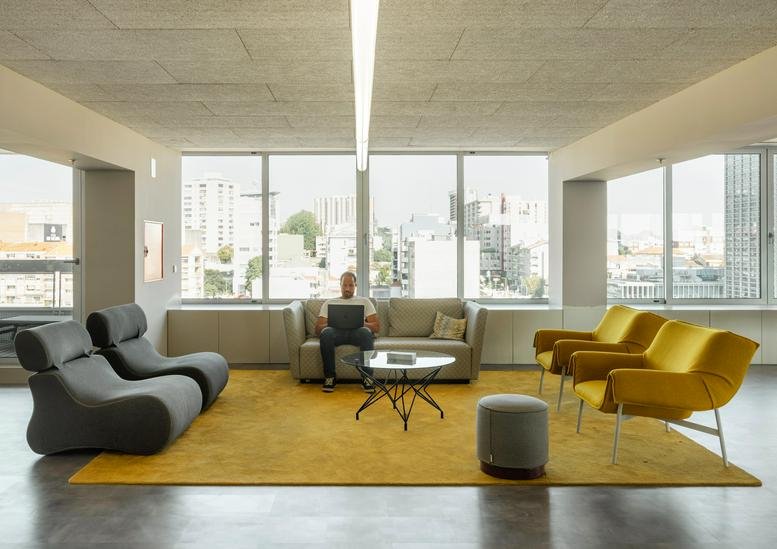 Modern communal lounge at R. de Azevedo Coutinho 39 with yellow rug, grey armchairs, and city views.