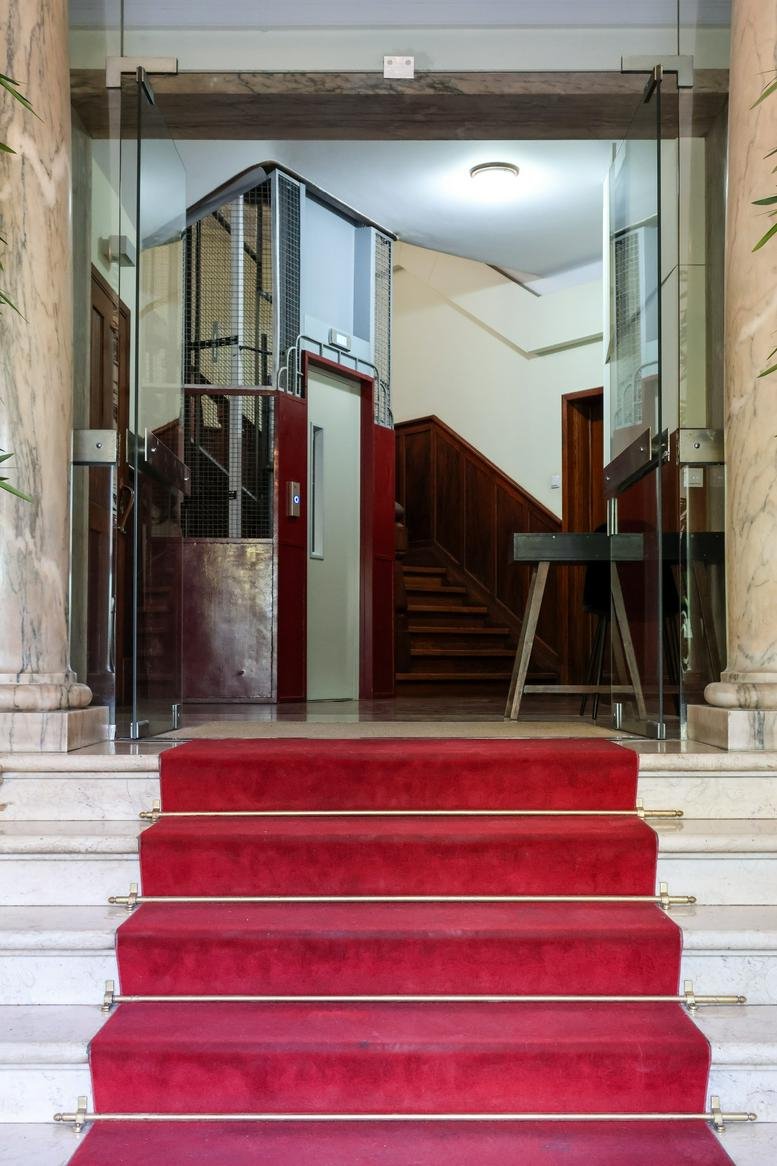 Grand entrance featuring marble stairs with a red carpet leading to glass doors and an elevator.
