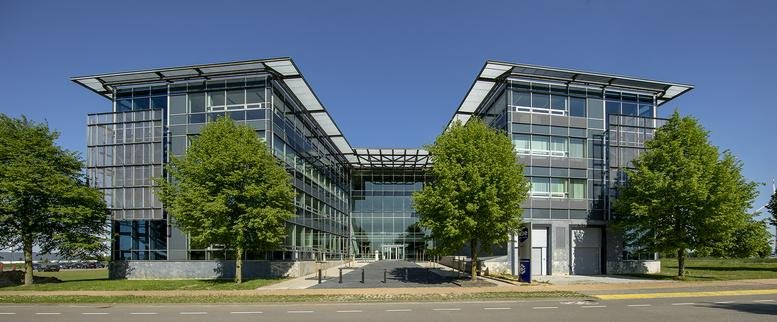 Front exterior of a four-story glass office building with large green trees and clear blue sky.