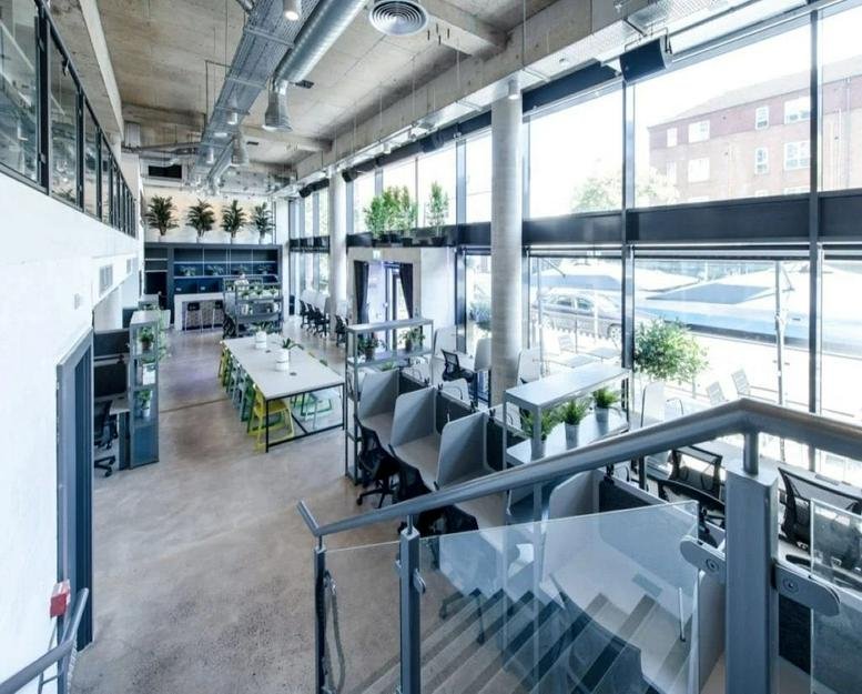 Spacious double-height coworking area with industrial ceilings and large glass windows.