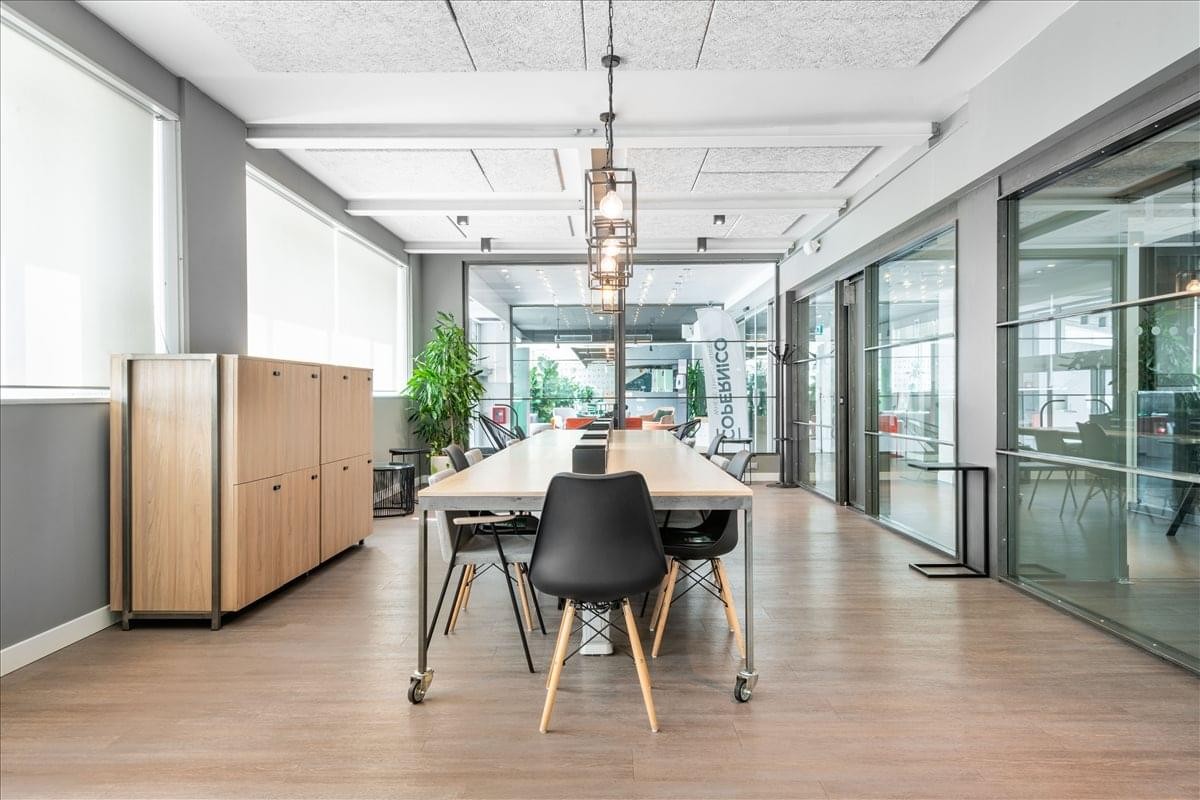 Open-plan office area at Viale Monza, 259/265 with a long wooden desk and glass-partitioned meeting rooms.