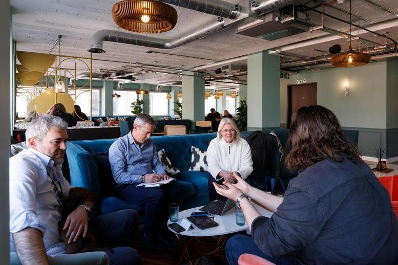 Bright communal lounge with blue velvet sofas, wooden light fixtures, and people working.